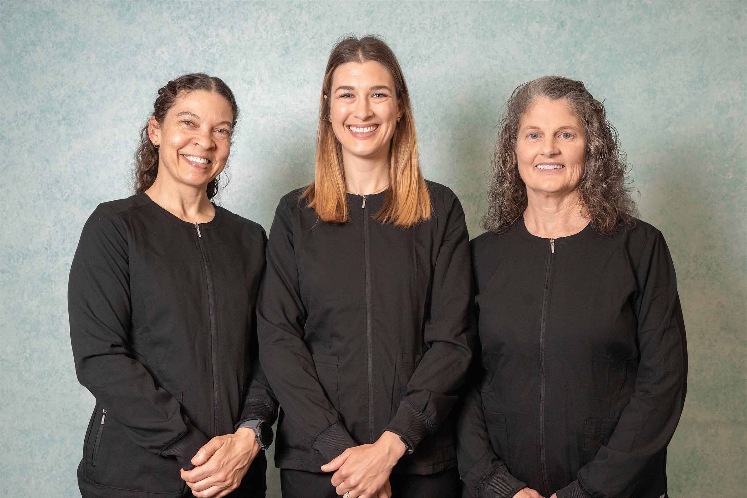 Three dental staff members standing side by side wearing black uniforms and smiling at the camera. The portrait shows a friendly clinic team posed against a neutral textured background.