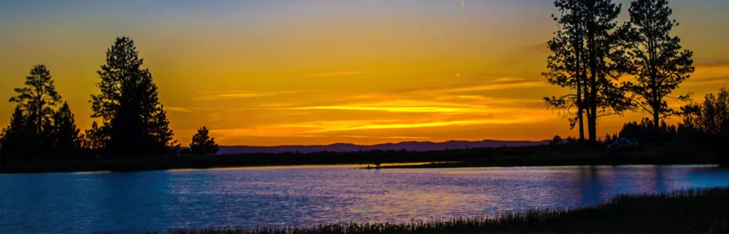 Augusta lake at sunset over a tranquil lake with golden-orange sky reflecting on calm water; silhouetted trees and distant hills frame the horizon. Peaceful, nature-rich scene evokes calm and reflection.