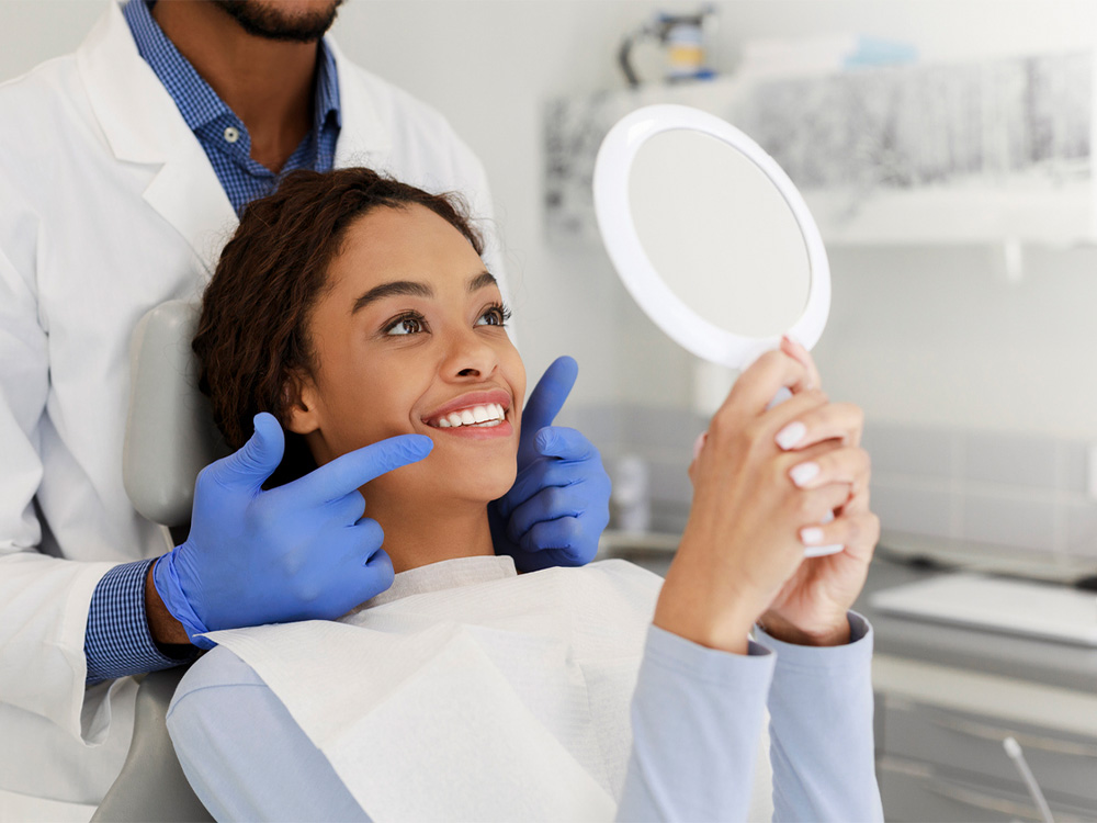 female patient smiling at her white teeth while looking in a mirror and the doctor behind her